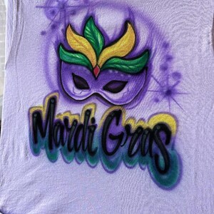 May include: A lilac t-shirt with an airbrushed Mardi Gras design. The design features a purple mask with yellow and green details, and the words "Mardi Gras" in a colourful, stylised font.