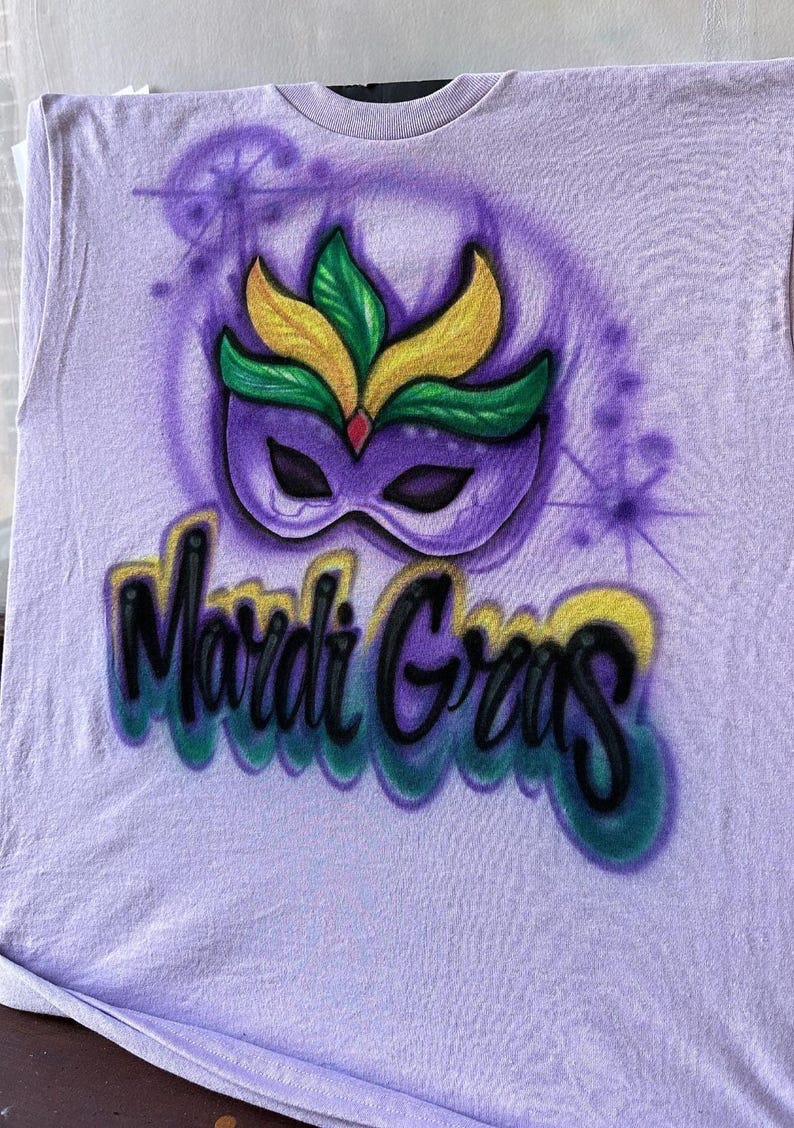 May include: A lilac t-shirt with an airbrushed Mardi Gras design. The design features a purple mask with yellow and green details, and the words "Mardi Gras" in a colourful, stylised font.