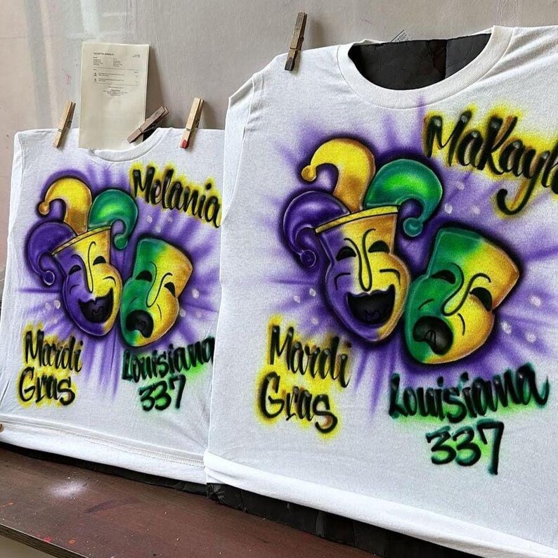 May include: White t-shirts with airbrushed Mardi Gras designs. Each shirt features a jester mask design in purple, green, and yellow, with the words "Mardi Gras" and "Louisiana 337". One shirt says "Melania" and the other says "Makayla".