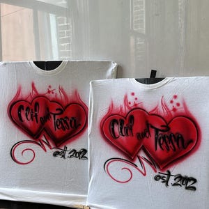 May include: Two white t-shirts with red and black airbrushed hearts. The hearts are intertwined and have the text "Cliff and Tessa est. 2012" written on them.