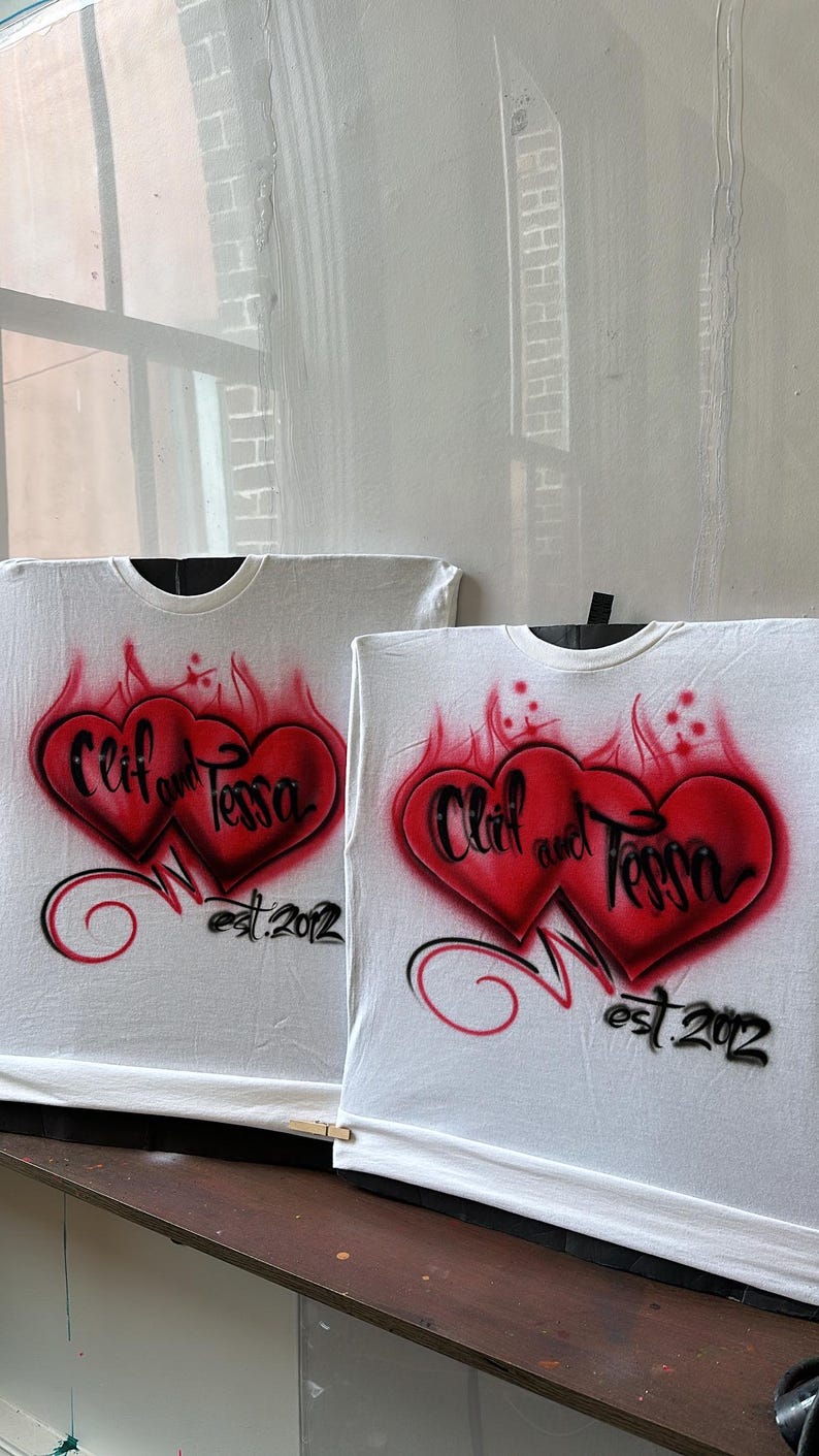 May include: Two white t-shirts with red and black airbrushed hearts. The hearts are intertwined and have the text "Cliff and Tessa est. 2012" written on them.