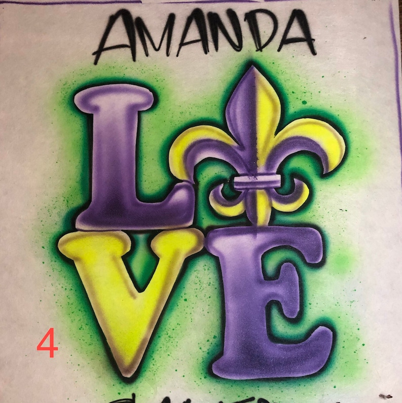 May include: Airbrushed design featuring the word "LOVE" in purple and yellow, with a fleur-de-lis symbol. The name "AMANDA" is above the word "LOVE". The background is green with a white border.