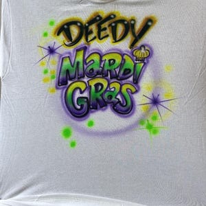 May include: White t-shirt with a colourful airbrushed design. The design features the words "DEEDY MARDI GRAS" in green, purple, and yellow, with starburst accents. A festive apparel item.