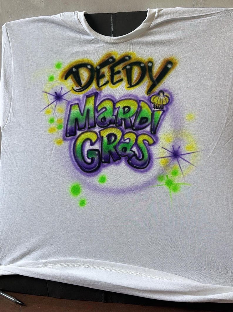 May include: White t-shirt with a colourful airbrushed design. The design features the words "DEEDY MARDI GRAS" in green, purple, and yellow, with starburst accents. A festive apparel item.