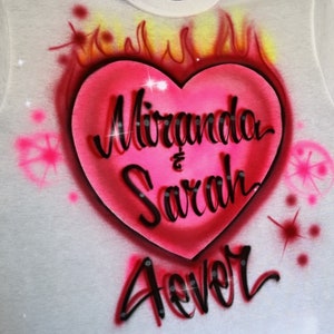 May include: White t-shirt with a pink heart design. The heart has flames around it and the text "Miranda & Sarah Forever" inside.