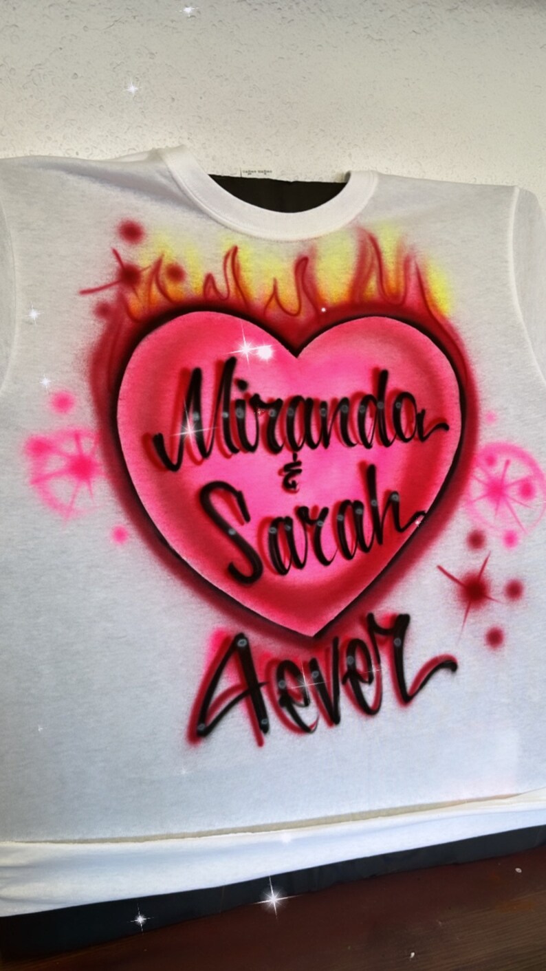 May include: White t-shirt with a pink heart design. The heart has flames around it and the text "Miranda & Sarah Forever" inside.