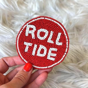 May include: A round, beaded coaster with the words "ROLL TIDE" in white letters on a red background. The coaster has a white beaded border and is held by a hand with red nail polish.