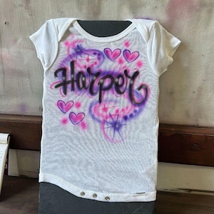 May include: White baby onesie with the name "Harper" in black script, surrounded by pink and purple airbrushed designs, including hearts and starbursts. The onesie has short sleeves and snap closures.