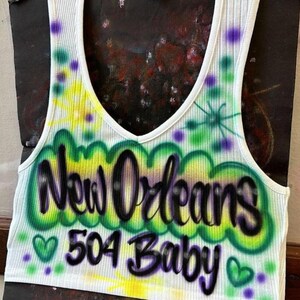 May include: White ribbed vest top with a V-neck. It features airbrushed lettering saying "New Orleans 504 Baby" in green and black, with green hearts and purple accents. The top has a colourful, airbrushed design.