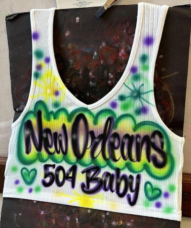 May include: White ribbed vest top with a V-neck. It features airbrushed lettering saying "New Orleans 504 Baby" in green and black, with green hearts and purple accents. The top has a colourful, airbrushed design.