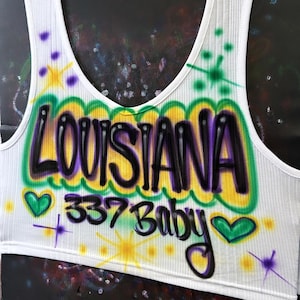May include: White ribbed vest top with an airbrushed design. The design features the word "LOUISIANA" in black and purple, outlined in yellow and green, with "337 Baby" below. Includes green hearts and starburst accents.