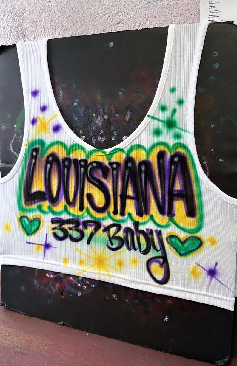 May include: White ribbed vest top with an airbrushed design. The design features the word "LOUISIANA" in black and purple, outlined in yellow and green, with "337 Baby" below. Includes green hearts and starburst accents.