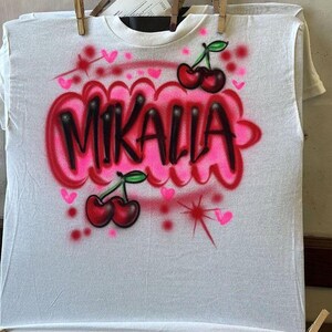 May include: White t-shirt with airbrushed design. The design features the name "MIKALLA" in black outlined in pink, with pink hearts and cherries. The shirt is held by wooden clothespins.