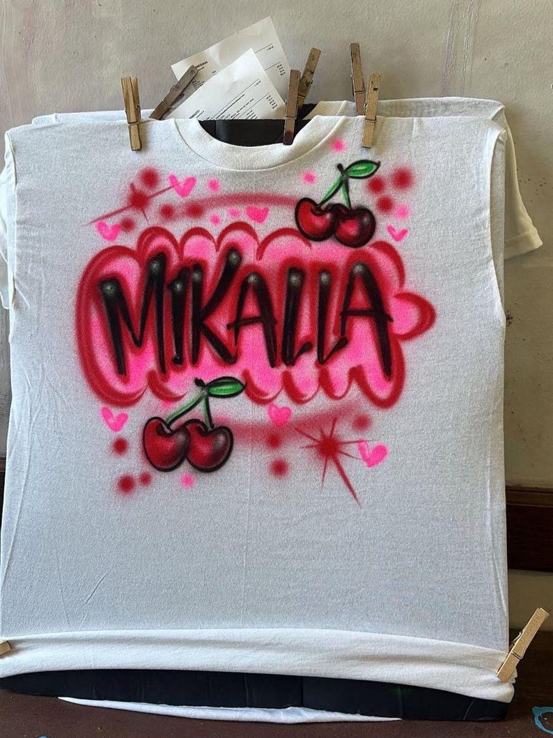 May include: White t-shirt with airbrushed design. The design features the name "MIKALLA" in black outlined in pink, with pink hearts and cherries. The shirt is held by wooden clothespins.