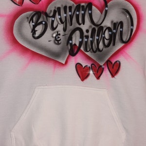 May include: White hoodie with a pink and grey heart design. The heart has the names "Brynn & Dillon" written in black with rhinestones. There are three smaller red hearts at the bottom of the design.