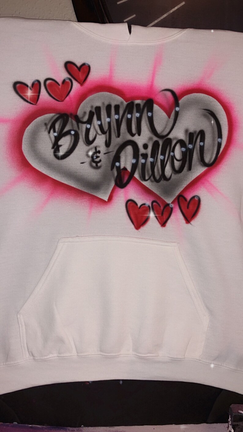 May include: White hoodie with a pink and grey heart design. The heart has the names "Brynn & Dillon" written in black with rhinestones. There are three smaller red hearts at the bottom of the design.