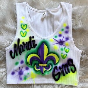 May include: White ribbed vest top with a V-neck. The front is decorated with airbrushed text reading "Mardi Gras" and a purple and yellow fleur-de-lis. Green, yellow, and purple accents complete the festive design.