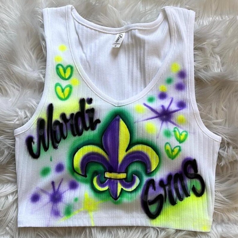 May include: White ribbed vest top with a V-neck. The front is decorated with airbrushed text reading "Mardi Gras" and a purple and yellow fleur-de-lis. Green, yellow, and purple accents complete the festive design.