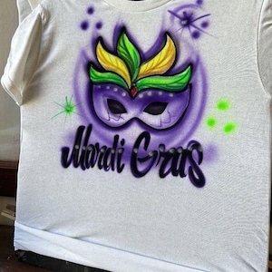 May include: White t-shirt featuring an airbrushed Mardi Gras design. The design includes a purple mask with green and yellow feathers, set against a purple background. The words "Mardi Gras" are written in a stylized font below the mask.