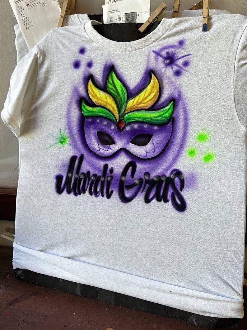 May include: White t-shirt featuring an airbrushed Mardi Gras design. The design includes a purple mask with green and yellow feathers, set against a purple background. The words "Mardi Gras" are written in a stylized font below the mask.