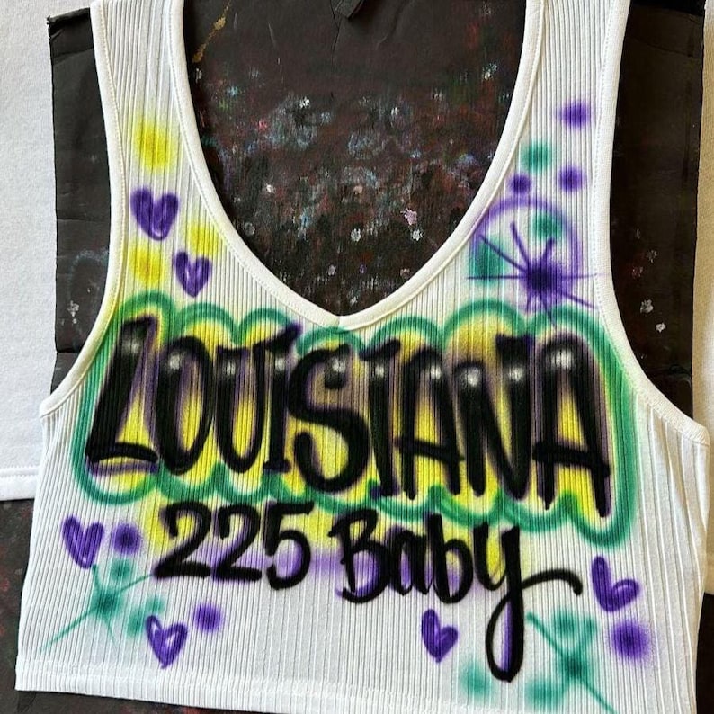 May include: White ribbed vest top featuring the word "LOUISIANA" in black graffiti-style lettering, outlined in green and yellow. The number "225" and the word "Baby" are below, with purple heart and star accents.