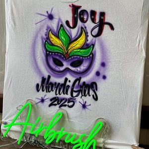 May include: White t-shirt with an airbrushed Mardi Gras design. The design features a purple mask with green and yellow accents, the word "Joy" in red, and "Mardi Gras 2025" in black. A neon green "Airbrush" sign is in front of the shirt.