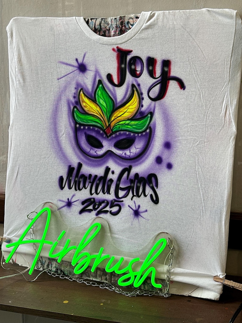 May include: White t-shirt with an airbrushed Mardi Gras design. The design features a purple mask with green and yellow accents, the word "Joy" in red, and "Mardi Gras 2025" in black. A neon green "Airbrush" sign is in front of the shirt.