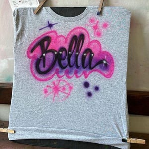 May include: A gray t-shirt with the name "Bella" airbrushed in a pink and purple cloud design. The shirt has additional pink and purple star and dot accents. The shirt is hanging with clothespins.
