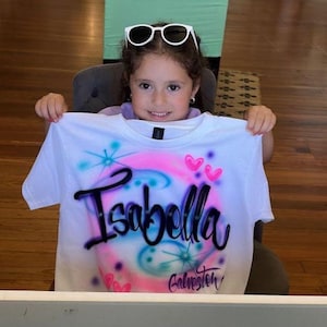 May include: White t-shirt with airbrushed design featuring the name "Isabella" in black script, surrounded by pink, blue, and purple swirls, hearts, and stars. The word "Galveston" is below the name. The shirt is held up by a person.