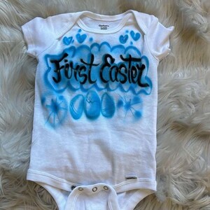 May include: White baby onesie with blue airbrushed design and the words "First Easter" in black. The design includes blue hearts and Easter eggs, perfect for a baby's first Easter celebration.