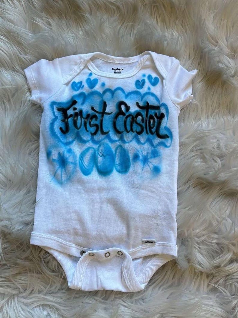 May include: White baby onesie with blue airbrushed design and the words "First Easter" in black. The design includes blue hearts and Easter eggs, perfect for a baby's first Easter celebration.