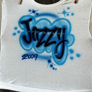 May include: White t-shirt with the word "Jazzy" in black, outlined in blue, and a blue cloud design. The year "2009" is below the word. The shirt is hanging from a clothesline with wooden clothespins.