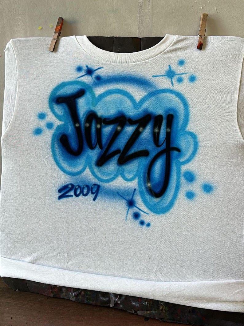 May include: White t-shirt with the word "Jazzy" in black, outlined in blue, and a blue cloud design. The year "2009" is below the word. The shirt is hanging from a clothesline with wooden clothespins.