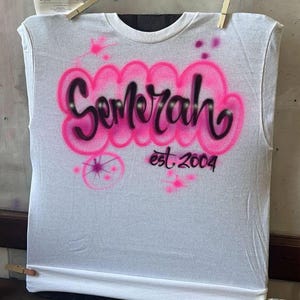 May include: White sleeveless t-shirt with pink and black airbrushed text that reads "Semerah" above "est. 2004". The design includes pink splatters and a starburst. The shirt is held in place with wooden clothespins.