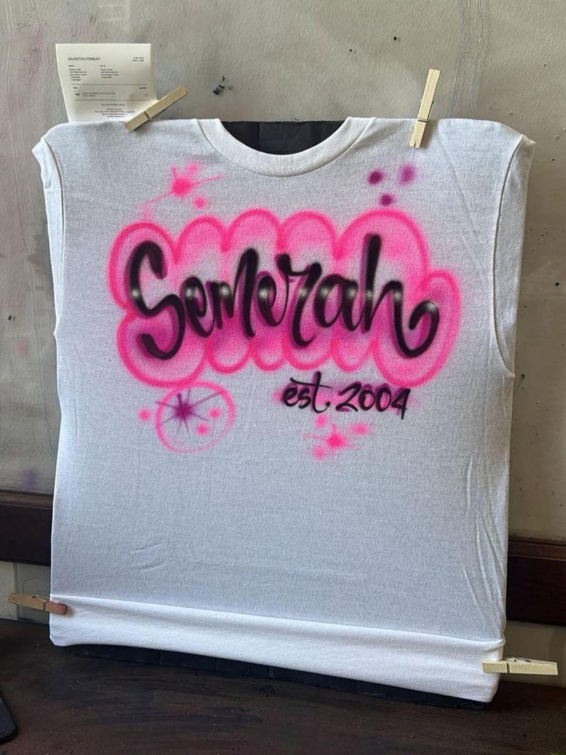 May include: White sleeveless t-shirt with pink and black airbrushed text that reads "Semerah" above "est. 2004". The design includes pink splatters and a starburst. The shirt is held in place with wooden clothespins.