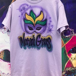May include: A lilac t-shirt with an airbrushed Mardi Gras mask design in green, yellow, and purple. The words "Mardi Gras" are written in a colourful, stylised font below the mask.