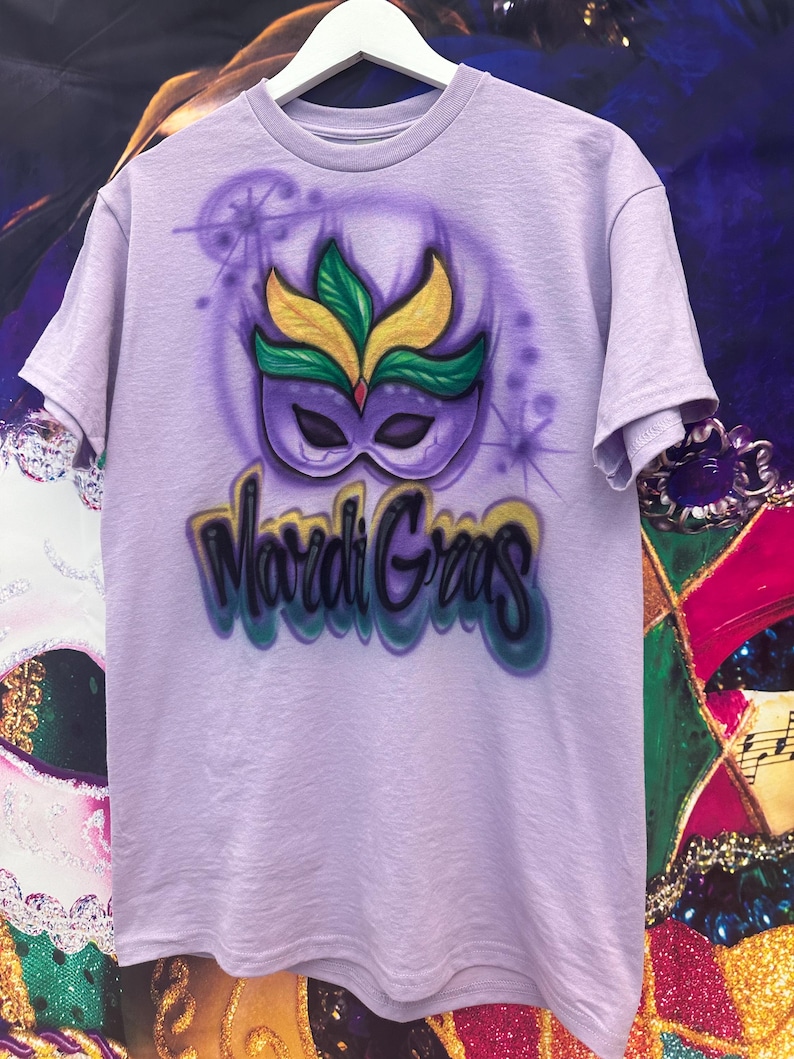 May include: A lilac t-shirt with an airbrushed Mardi Gras mask design in green, yellow, and purple. The words "Mardi Gras" are written in a colourful, stylised font below the mask.