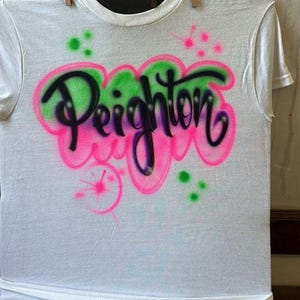 May include: White t-shirt with the name "Peighton" airbrushed in black, outlined with pink and green. The design includes pink and green paint splatters. The shirt is hanging with wooden clothespins.