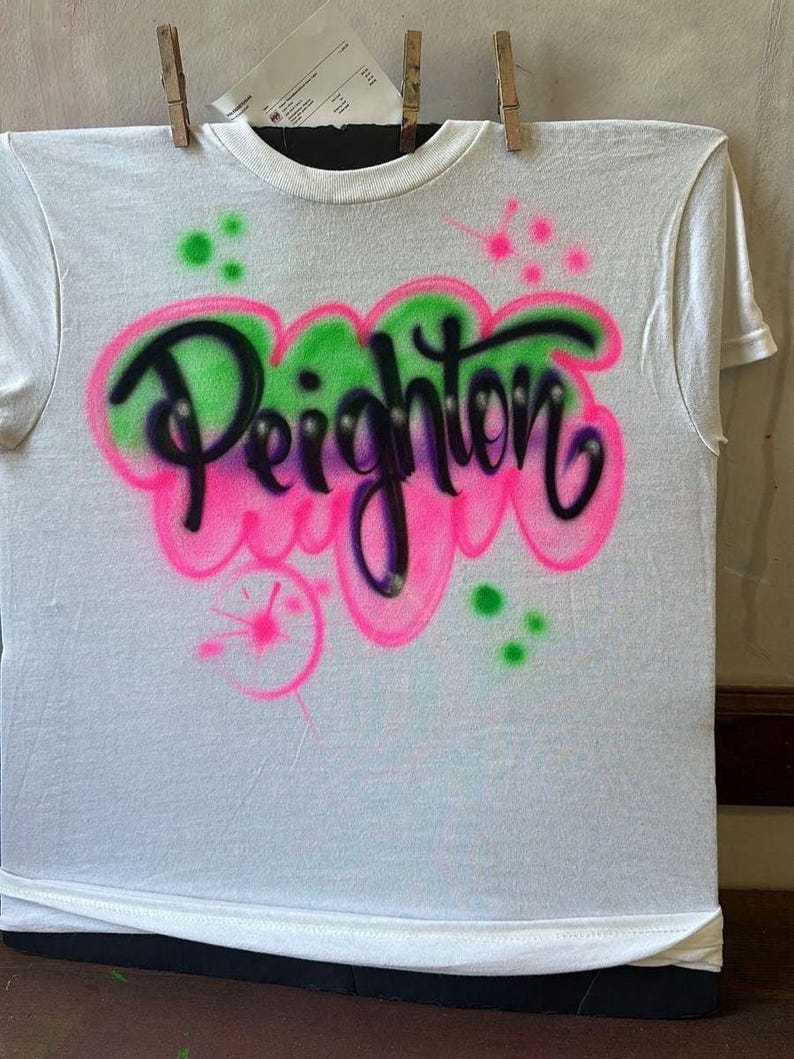 May include: White t-shirt with the name "Peighton" airbrushed in black, outlined with pink and green. The design includes pink and green paint splatters. The shirt is hanging with wooden clothespins.