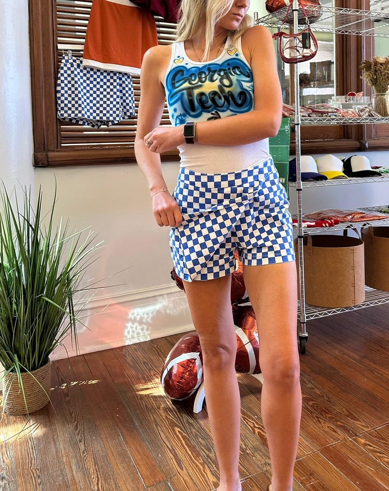 May include: A white vest top with "Georgia Tech" in blue and yellow lettering, paired with blue and white chequered shorts. A matching outfit is hanging in the background. The outfit is casual and sporty.