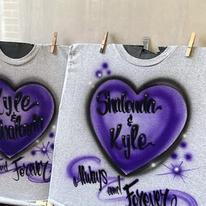May include: Two gray t-shirts with purple airbrushed hearts. The hearts have the names "Shalonda & Kyle" and "Kyle & Shalonda" written inside. The shirts also have the text "Always and Forever" written below the hearts.