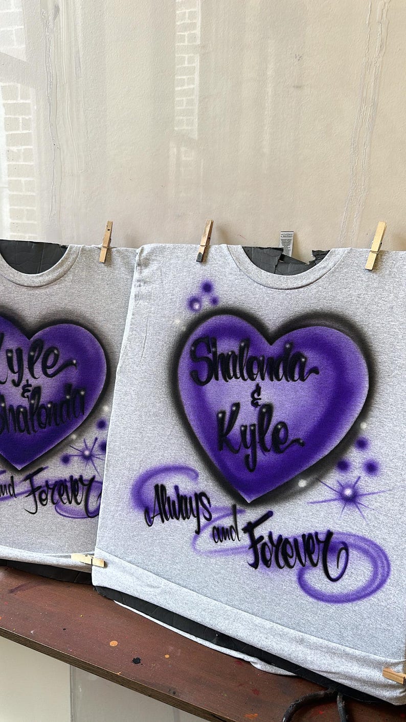 May include: Two gray t-shirts with purple airbrushed hearts. The hearts have the names "Shalonda & Kyle" and "Kyle & Shalonda" written inside. The shirts also have the text "Always and Forever" written below the hearts.