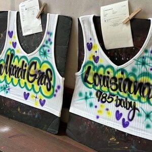 May include: Two white ribbed tank tops with a V-neck design. One top features the text "Mardi Gras" in black and yellow, with green and purple accents. The other top says "Louisiana 985 baby" in black, with green, yellow, and purple accents.