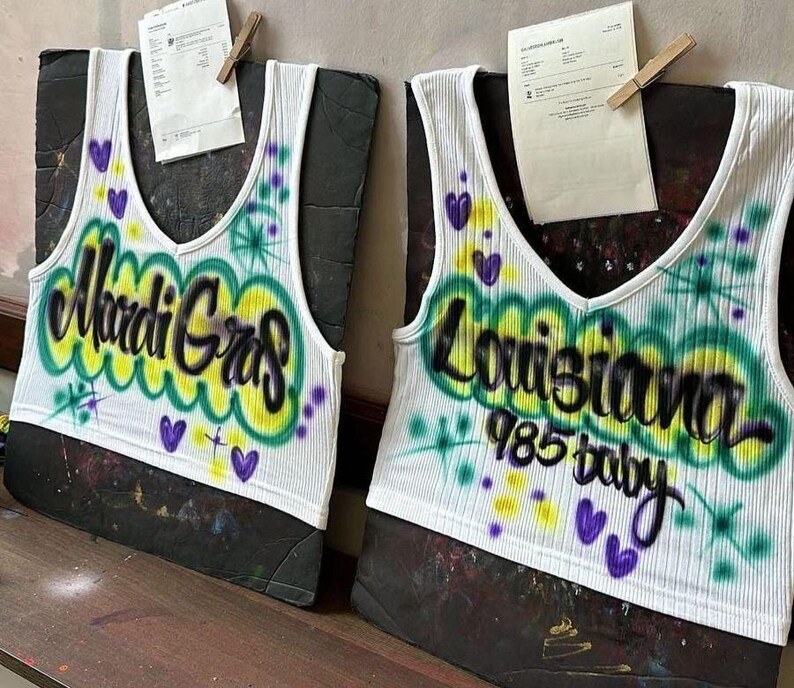 May include: Two white ribbed tank tops with a V-neck design. One top features the text "Mardi Gras" in black and yellow, with green and purple accents. The other top says "Louisiana 985 baby" in black, with green, yellow, and purple accents.