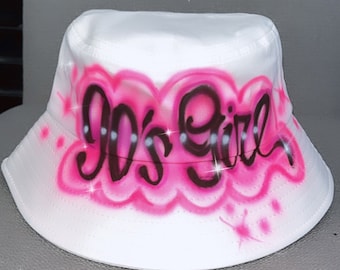 May include: White bucket hat with pink and black spray paint design that says "90's Girl".