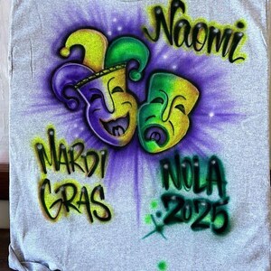 May include: Grey t-shirt with an airbrushed Mardi Gras design. It features two smiling masks in yellow, green, and purple, with the name "Naomi" above. The words "Mardi Gras" and "NOLA 2025" are also displayed.