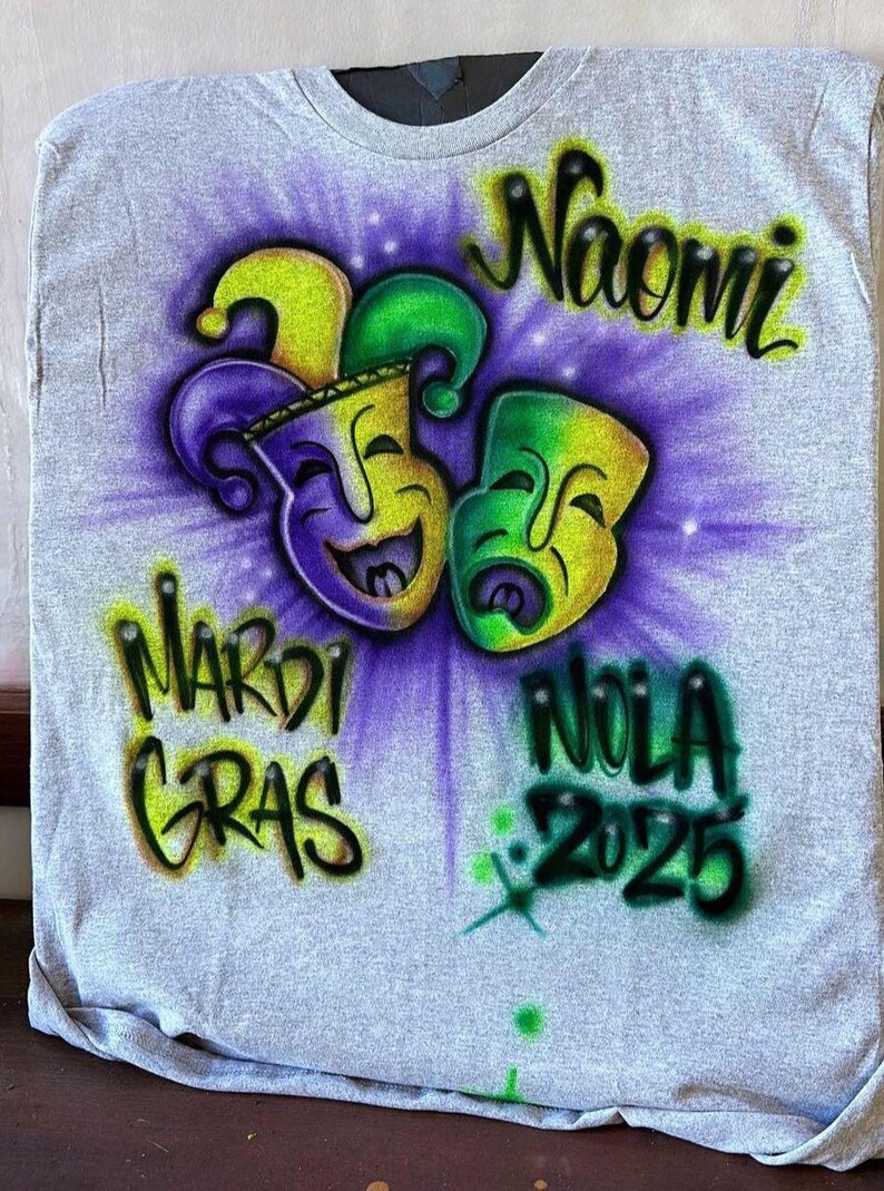 May include: Grey t-shirt with an airbrushed Mardi Gras design. It features two smiling masks in yellow, green, and purple, with the name "Naomi" above. The words "Mardi Gras" and "NOLA 2025" are also displayed.