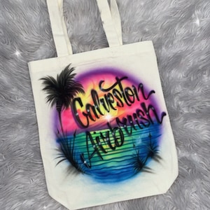 May include: A white canvas tote bag with a colorful airbrushed design of a sunset over the ocean with palm trees and the text "Galveston Airbrush".