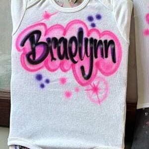 May include: White baby onesie with the name "Braelynn" airbrushed in black and pink. The name is surrounded by pink clouds and accented with purple and pink starbursts. A cute and personalized baby garment.