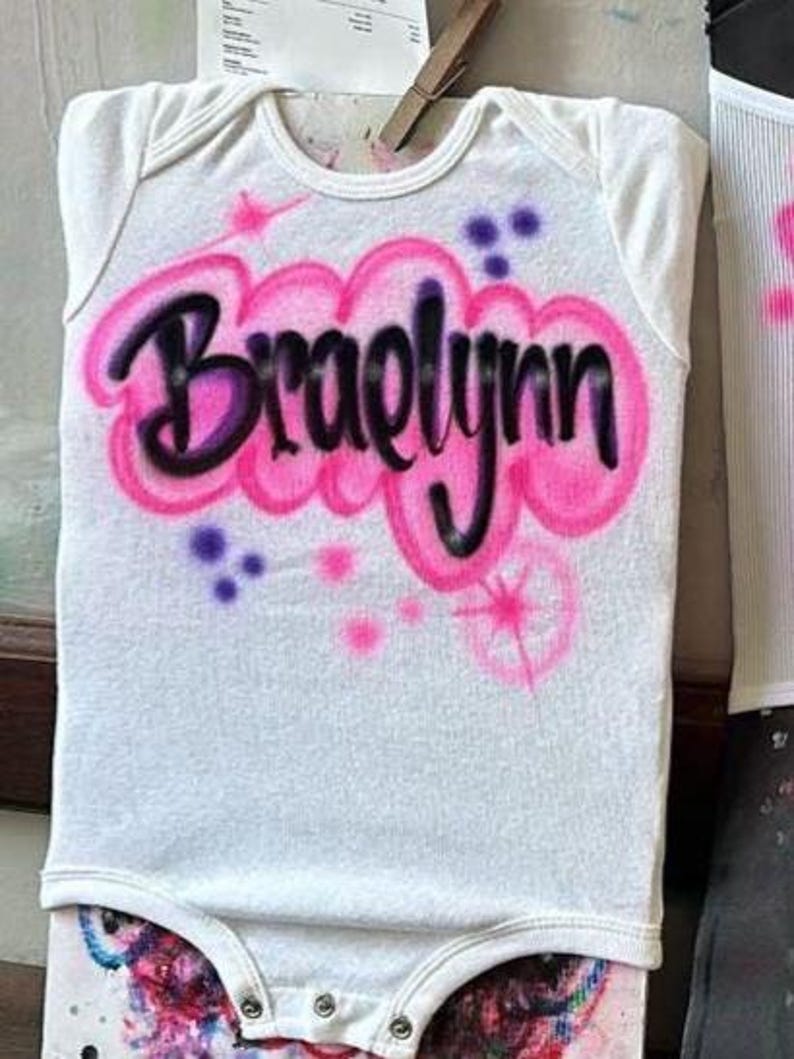 May include: White baby onesie with the name "Braelynn" airbrushed in black and pink. The name is surrounded by pink clouds and accented with purple and pink starbursts. A cute and personalized baby garment.
